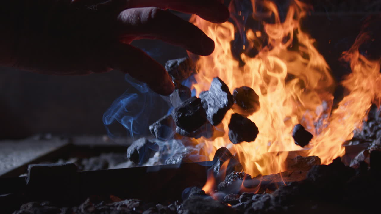 Hand throws extra coals on blacksmith's forge with hot raging orange flames