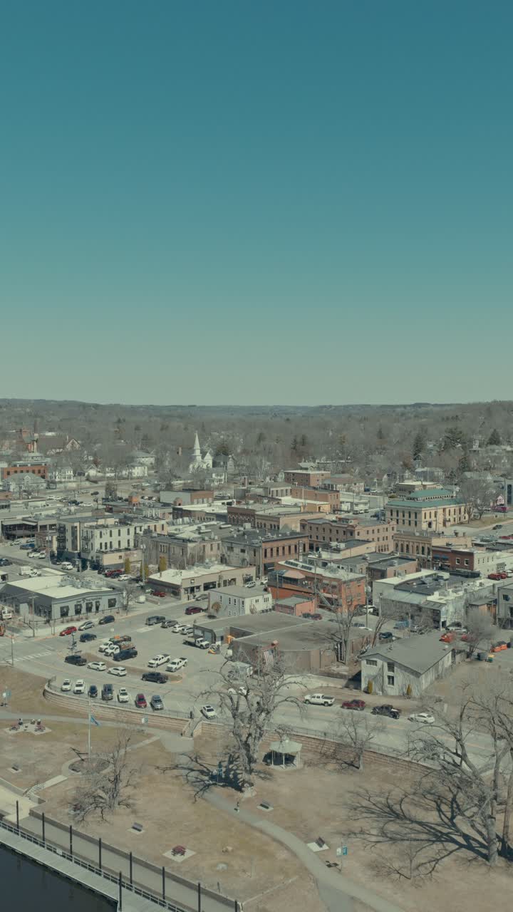 Volo aereo verticale in avanti da sopra il fiume St. Croix verso il centro di Hudson, Wisconsin, che rivela la passeggiata sul lungofiume, edifici storici in mattoni, il campanile di una chiesa bianca e scogliere boscose.