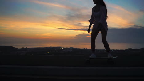 in a slow-motion scene, a stylish young skateboarder rides her board in shorts at sunset along a mountain road, offering a stunning mountain view