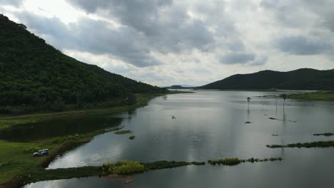 aerial footage sliding towards the left revealing the beautiful lake and mountains then a truck and some people getting ready to leave after fishing for the whole day; saraburi, thailand