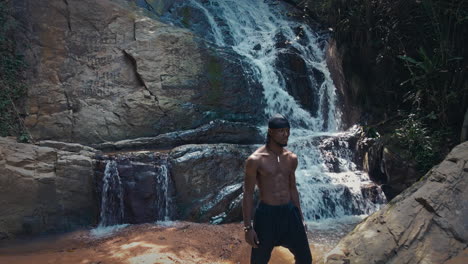 drone forward moving shot of a black male standing on the rock in front of jungle waterfall surrounded by lush green vegetation at daytime