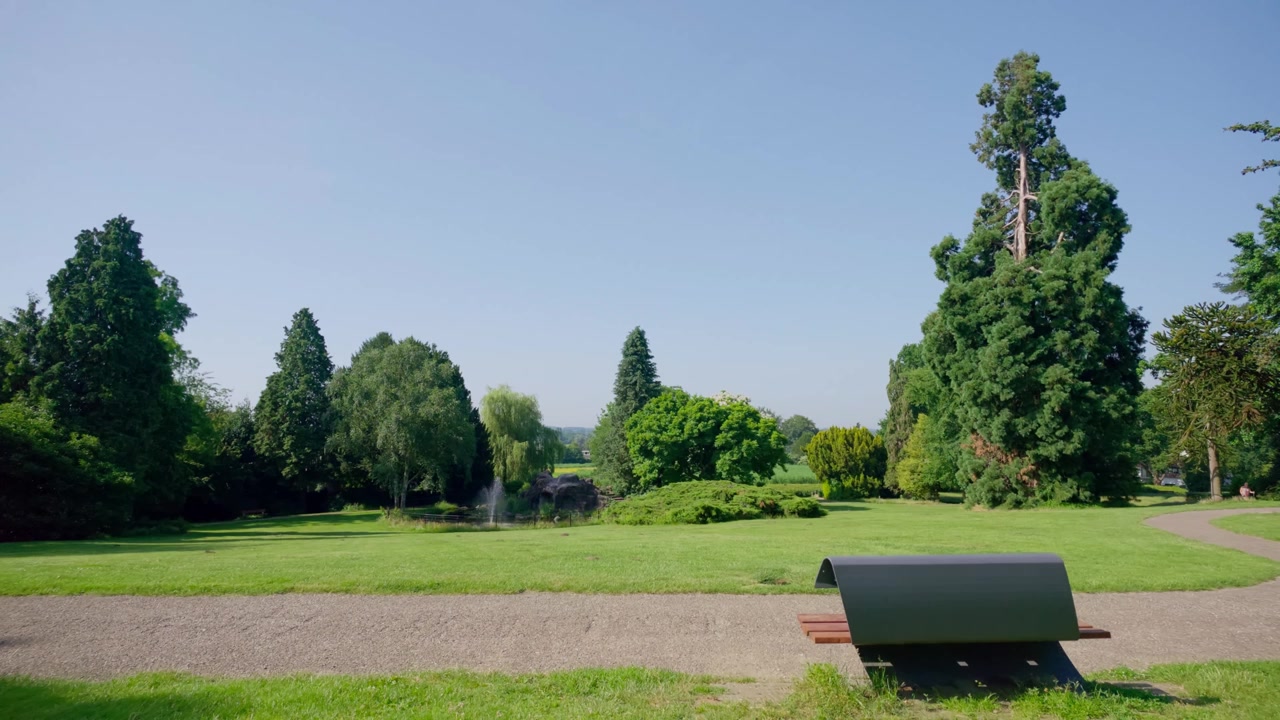 Weitläufige Ansicht eines ruhigen Parks mit einem gewundenen Kiesweg, einem friedlichen Teich mit einem sanften Springbrunnen und vielfältigen alten Bäumen. Eine einzelne Parkbank bietet einen Ruheplatz inmitten der natürlichen Schönheit.