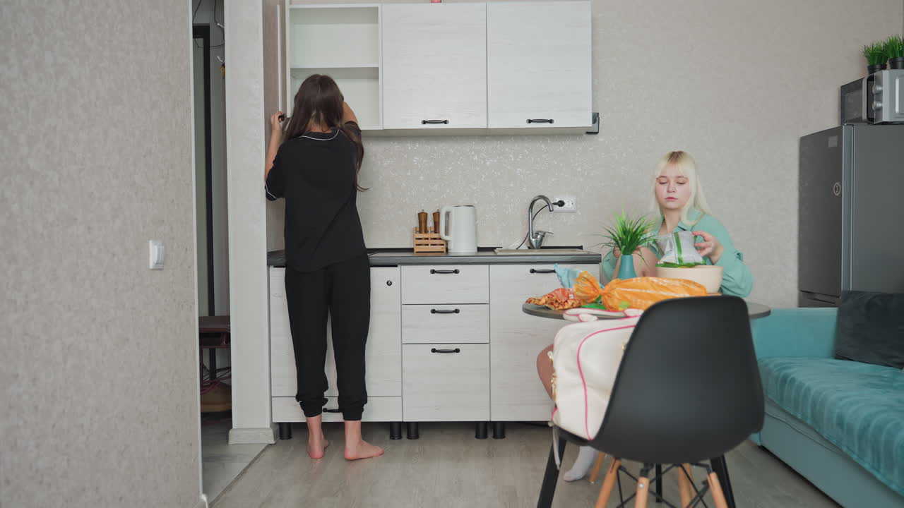 woman in black pajamas hands bowl to friend seated at table who adds cheese into bowl in cozy modern kitchen during morning routine, bright decor, and warm atmosphere of friendship