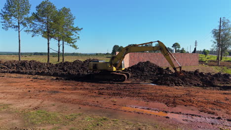 yellow excavator digging red dirt at construction site near trees and built wall.