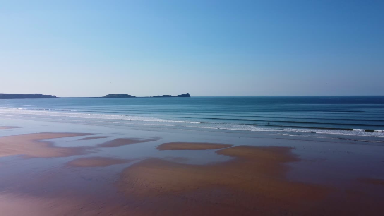 Descending Aerial View of Sea Waves Rolling into Rhossili Bay Beach with Worm's Head Landmark Silhouetted on Horizon 4K