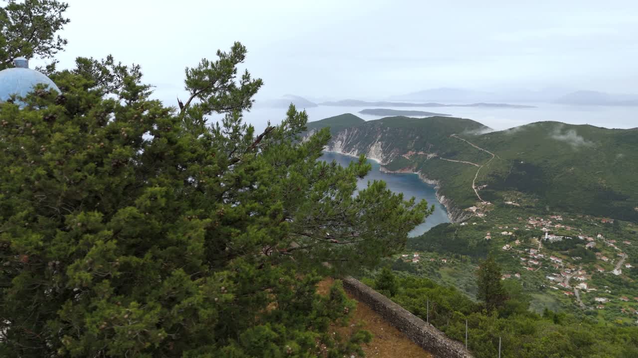 Aerial drone view from the Monastery of Panagia Eleousa atop a mountain in Ithaca, Greece, overlooking rolling landscapes, small villages, and lush green forests.