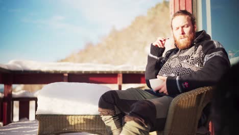 Bärtiger-Mann-Mit-Einer-Schüssel,-Der-Schokolade-Isst,-Sitzt-Mit-Seinem-Hund-Auf-Dem-Balkon-Einer-Hütte-An-Einem-Wintertag-Im-Trondheim-Fjord,-Norwegen