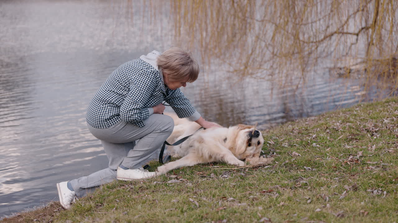 una mujer mayor jugando con su golden retriever junto a un lago.