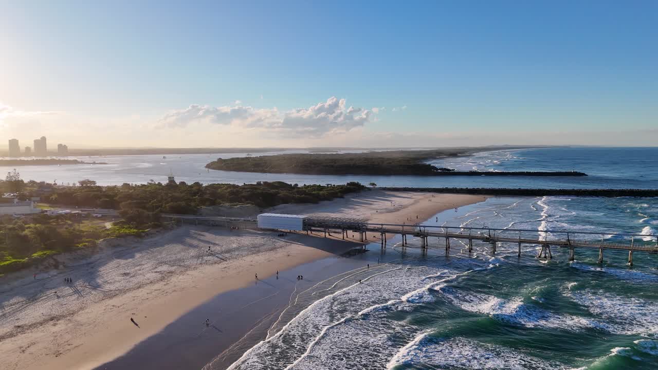 Drone footage captures the serene Gold Coast beach and jetty at sunset, highlighting waves and coastal landscape