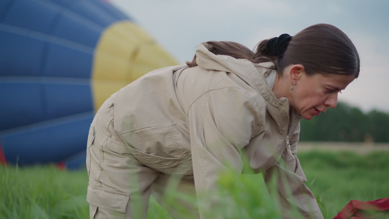 lady adjusting vibrant balloon envelope fabric on grassy field during prep setup bending forward in durable jumpsuit and gloves near partially inflated hot air balloon canopy under cloudy sky at dusk