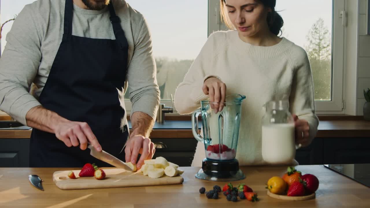 Un uomo in grembiule affetta fragole e una donna versa il latte nel frullatore a casa per preparare un frullato