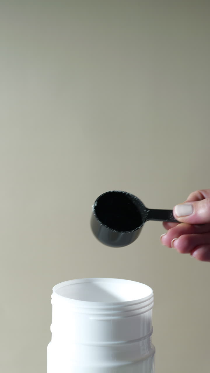 hand pours a scoop of white yellow protein powder—vanilla or banana flavored—into a clean white cup shaker, powder gently falling against a soft neutral background in a minimal, health-focused setup