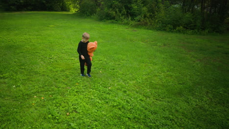 young child in black outfit holding large orange teddy bear while walking slowly and thoughtfully through bright grassy field surrounded by lush green trees under natural daylight