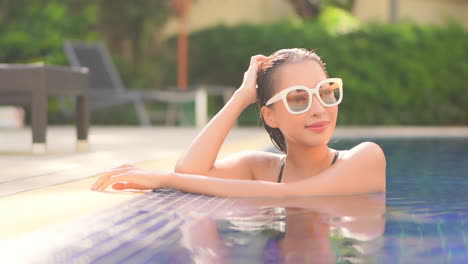 a close-up young attractive woman wearing sunglasses in a swimming pool leans on the edge of the pool turns to look around