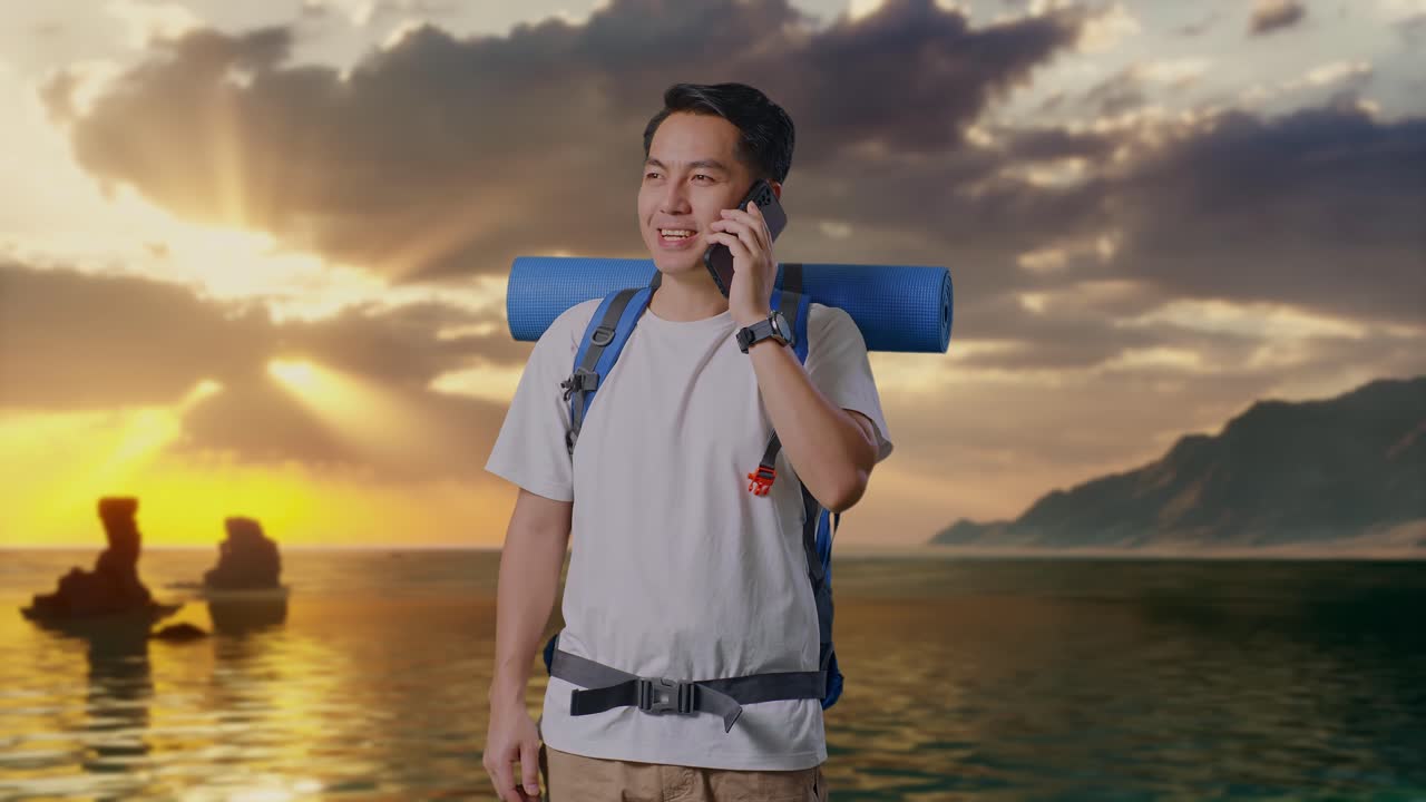 登山用バックパックを背負い、ビーチでスマートフォンで笑顔で話すアジア人男性ハイカー