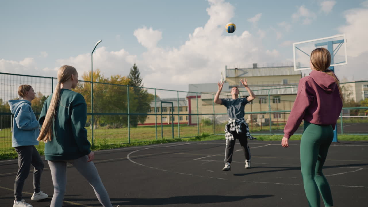 3명의 여성이 야외 코트에서 공을 던지는 배구 훈련을 주도하고 팀워크와 연습을 보여주는 장면, 건물, 울타리 및 농구 코트의 배경을 포함합니다.