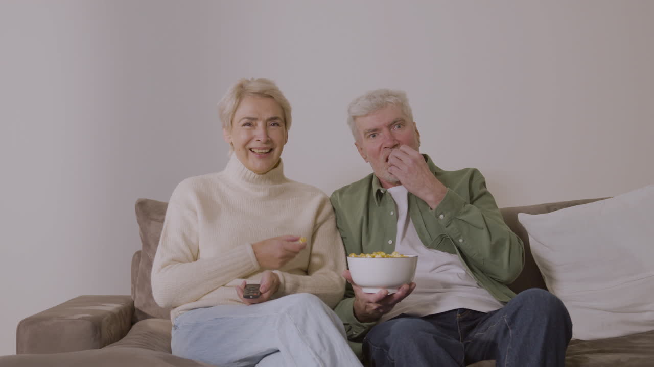Couple de seniors mangeant du pop-corn au caramel et regardant la télévision assis sur un canapé à la maison