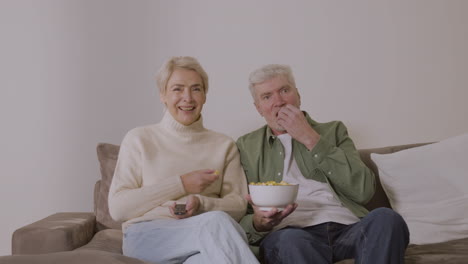 senior couple eating caramel popcorn and watching tv while sitting on sofa at home