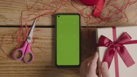 overhead shot of man gift wrapping romantic valentines present of perfume in box next to green screen mobile phone 1