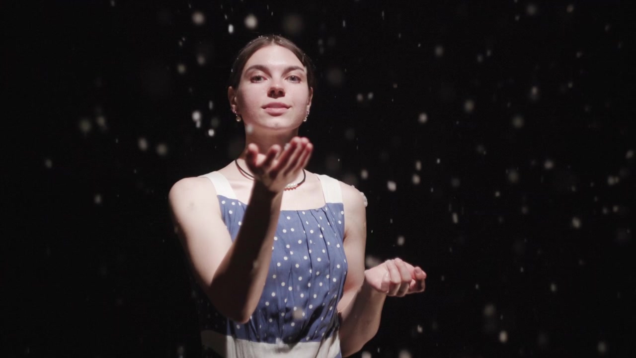 A young woman stands against a dark background with falling droplets, holding her hands raised in a gentle pose under soft studio lighting with a moody