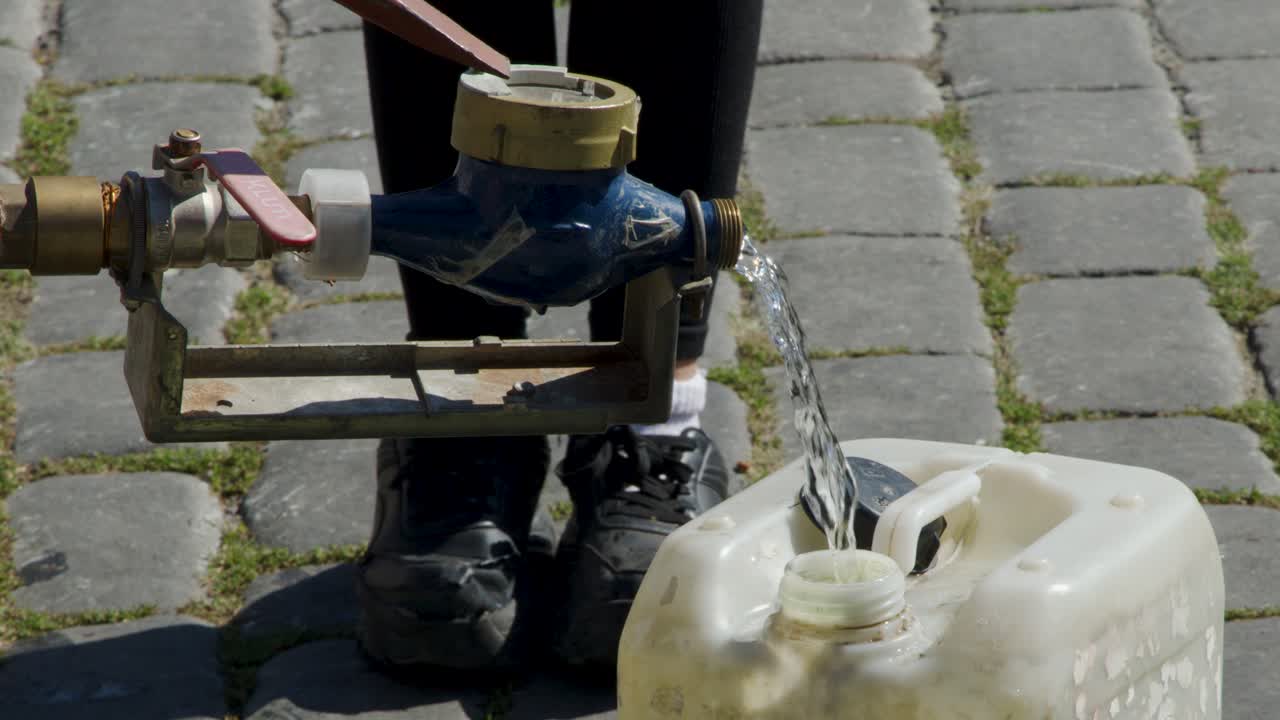Person fills large plastic bottle from metal street tap on cobblestone pavement in daylight