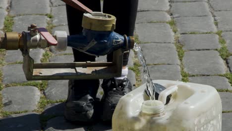 person fills large plastic bottle from metal street tap on cobblestone pavement in daylight