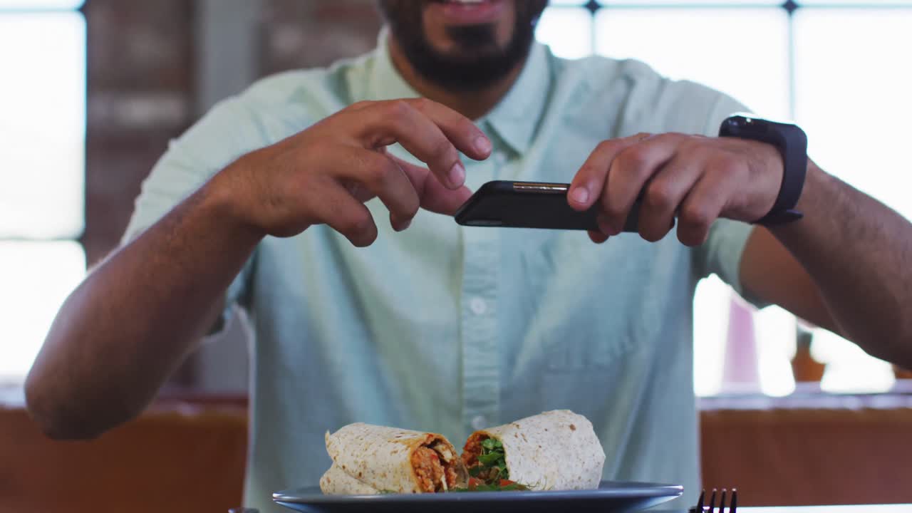 feliz hombre de raza mixta sentado en una cafetería tomando una foto de su sándwich con un teléfono inteligente y sonriendo