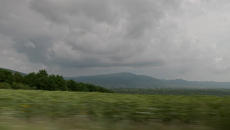 Side-view-of-the-mountains-from-the-moving-car-on-a-cloudy-day,-with-farmland-in-the-foreground