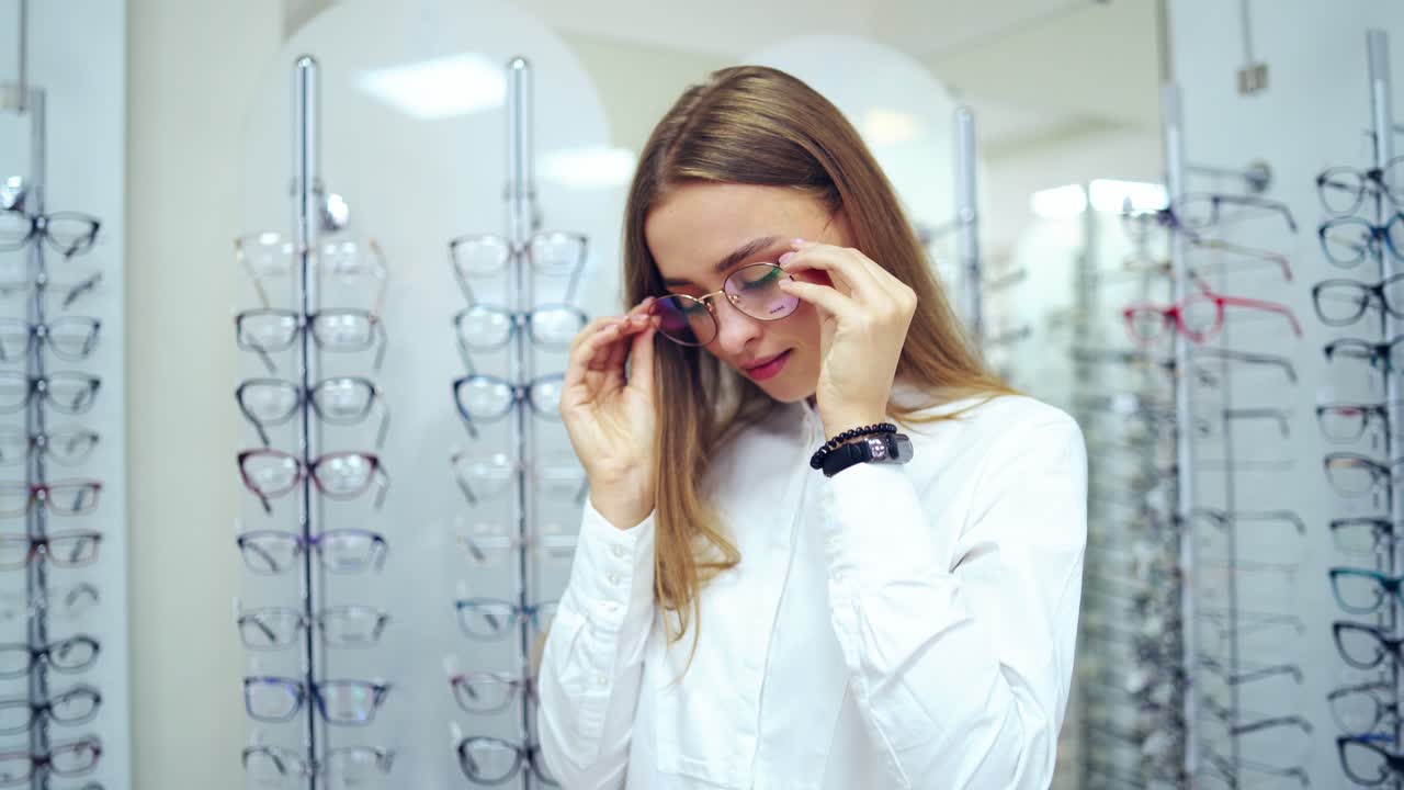 Fashionable eyeglasses in female's hands. Attractive young woman putting on new spectacles and smiling on camera at optics.