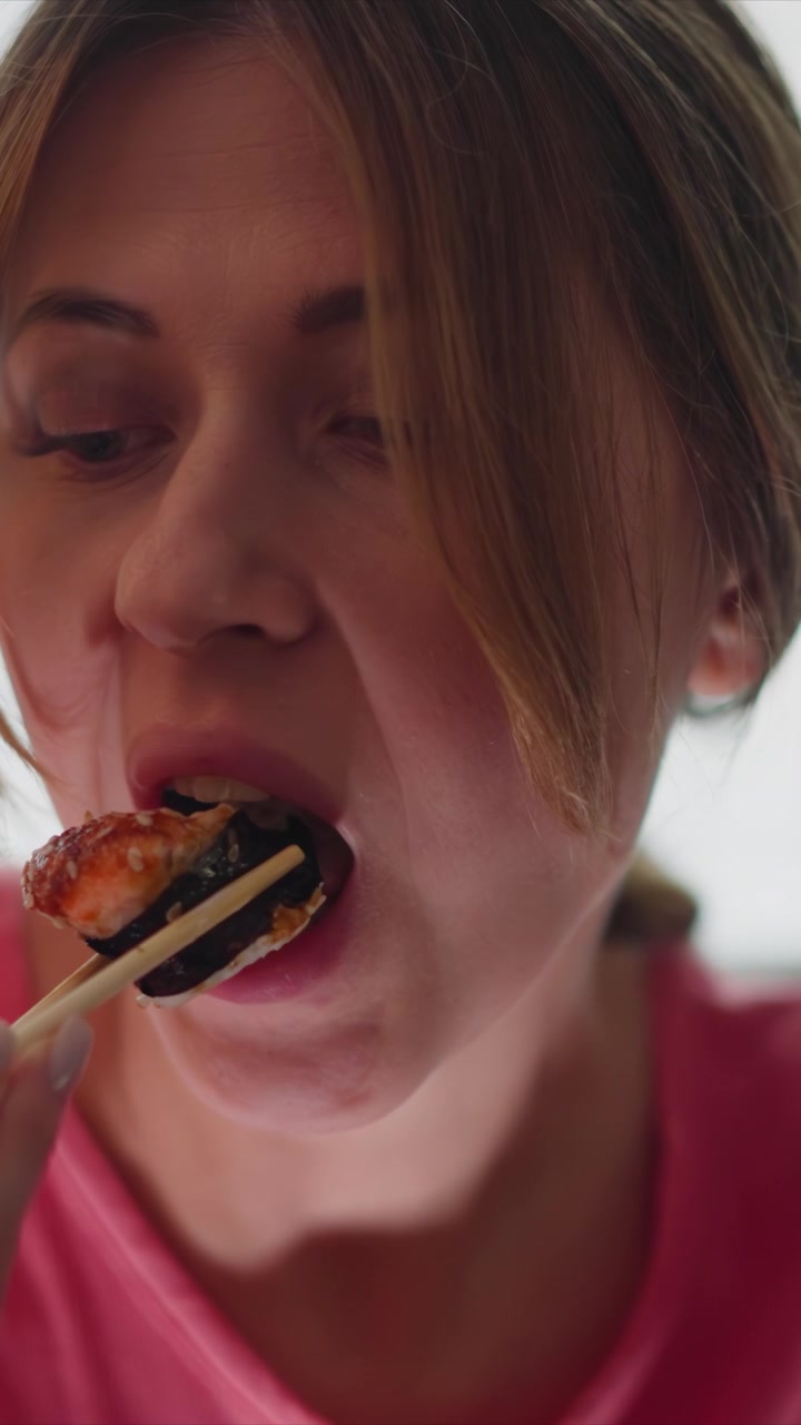 Close up of woman using chopsticks to eat sushi. scene focuses on action and texture details with soft lighting highlighting her pink shirt and food. Background remains out of focus