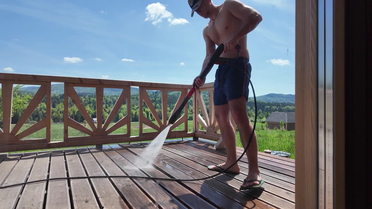 Hombre realiza mantenimiento exterior de casa lavando a presión una terraza en una cabaña rústica rodeada de campo verde, bosque y montañas.