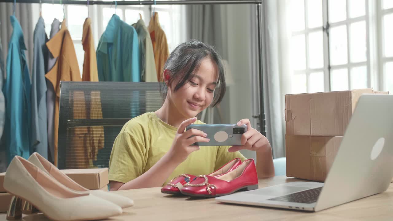 joven asiática vendedora en línea tomando fotos con teléfono móvil mientras usa la computadora para vender ropa en casa
