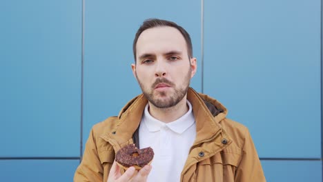 hungry man with a mustache and a beard eats a chocolate donut against a blue background