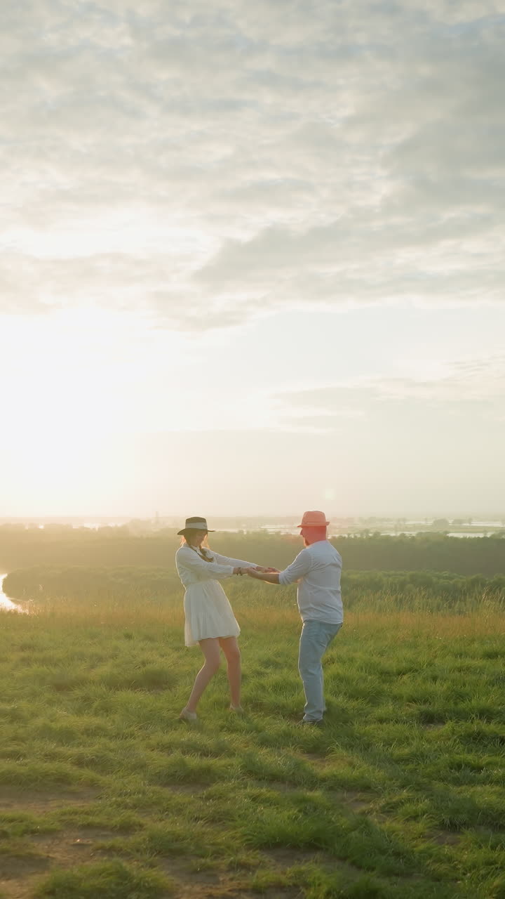한 사랑하는 부부는 평온한 호수 의 잔디 언덕에서 해가 지는 동안 기게 회전합니다. 남편은  셔츠와 모자를 입고 색 드레스를 입은 아내를 포옹하여 순수한 로맨스의 순간을 포착합니다.