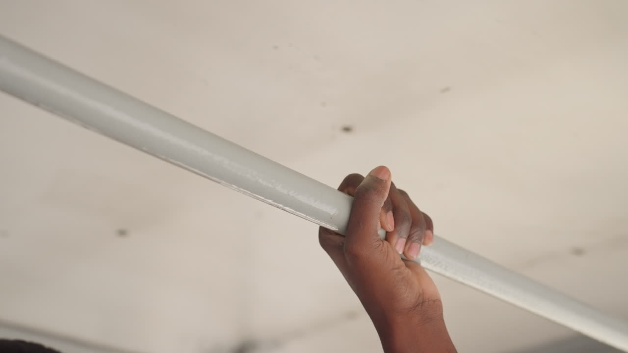 Mano nera che afferra un palo della metropolitana, polsino di maglione arancione visibile, primo piano del palmo e delle dita che afferrano la barra di metallo, illuminazione fluorescente e sfocatura di movimento sottile suggeriscono un vagone in movimento, pendolare che si mantiene.
