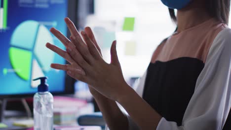 Mid-section-of-asian-woman-wearing-face-mask-sanitizing-her-hands-at-modern-office