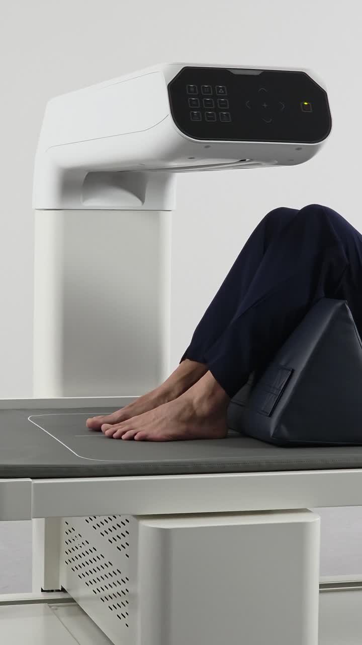 A vertical tracking shot of a male patient lying on a table with his knees supported by a positioning cushion, doing a spinal bone density DEXA scan in a modern hospital.