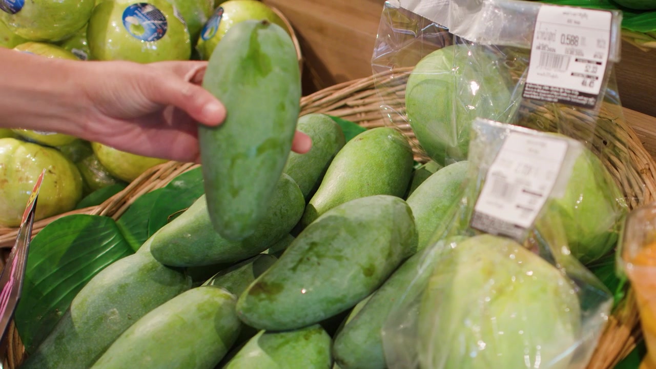 Un primo piano mostra una mano che raccoglie e ispeziona manghi verdi in un supermercato.
