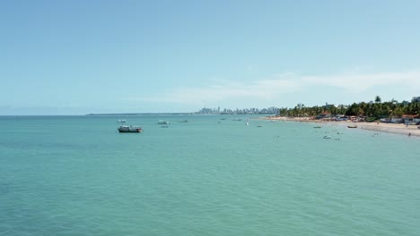 trucking left aerial wide shot of the tropical bessa beach in the coastal capital city of joao pessoa, paraiba, brazil with people enjoying the ocean and fishing boats at shore on a sunny summer day