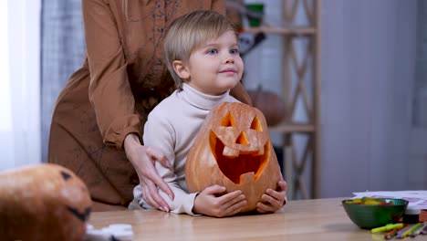 the boy sits at a table looks ahead and hugs a large orange pumpkin - the boy sits at a table in the room looks ahead and hugs a large orange pumpkin jack o lantern. the child begins to be amazed at something joyfully, and his mother stands behind him and strokes his hands. preparing for halloween.