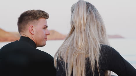 Rear-View-Of-Couple-Wearing-Wetsuits-Sitting-On-Beach-Talking-And-Looking-Out-To-Sea-Together