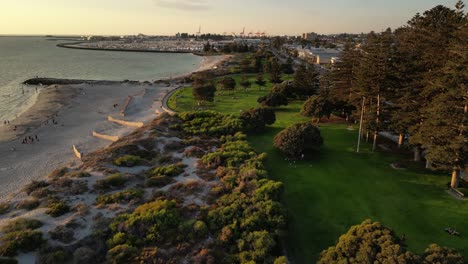 Antena-Que-Establece-Un-Vuelo-De-Drones-Sobre-Las-Dunas,-La-Playa-De-Arena-Y-El-Parque-De-La-Playa-Del-Sur-En-Fremantle,-Australia-Occidental---Puerto-Del-Puerto-En-Segundo-Plano-Durante-La-Puesta-De-Sol-Dorada