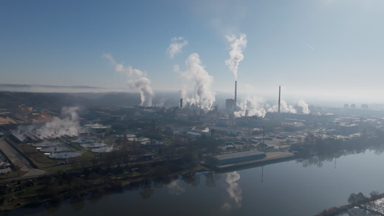 Panorama luas dari drone industri di tepi sungai di Štětí, menangkap cerobong asap, atap pabrik, kolam pengolahan, dan koridor sungai di bawah sinar matahari yang kabur