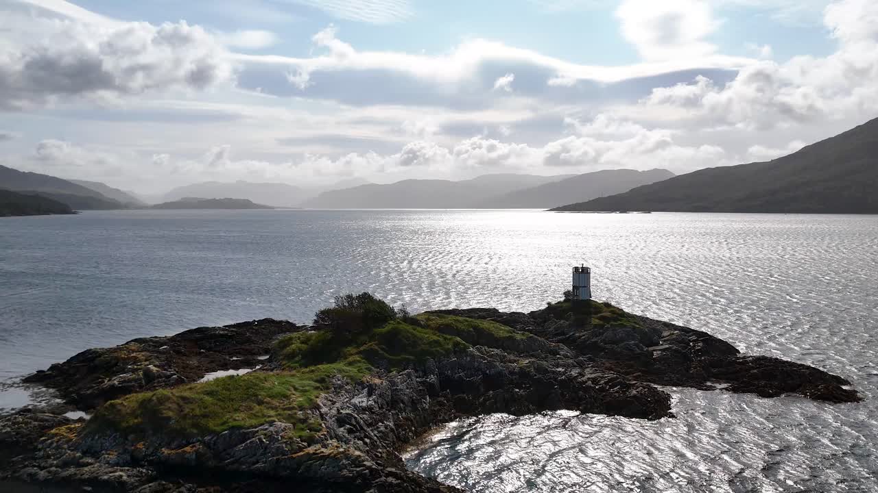 Drone glides toward a small island lighthouse on Loch Alsh, Isle of Skye, under dramatic clouds and bright backlighting, revealing rugged Scottish landscape