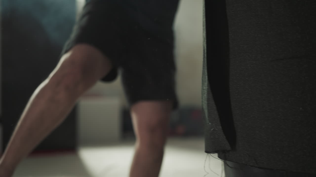 활기찬 남성 운동 선수는 키크박싱을 위해 지하실에서 벌거벗은 다리로 매달린 싱 가방을 때립니다.