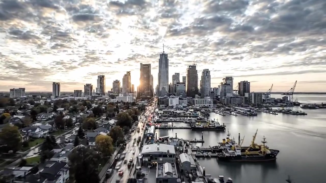 Una dinamica scena urbana: ciclisti e auto si muovono attraverso un paesaggio stradale cittadino circondati da edifici moderni e infrastrutture in una giornata di pioggia.