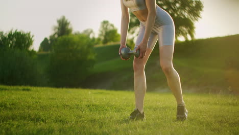 una mujer joven en ropa deportiva se inclina hacia adelante con pesas en la naturaleza en un parque al atardecer. entrenamiento. trabajar en un cuerpo hermoso por la mañana o al atarrear el sol