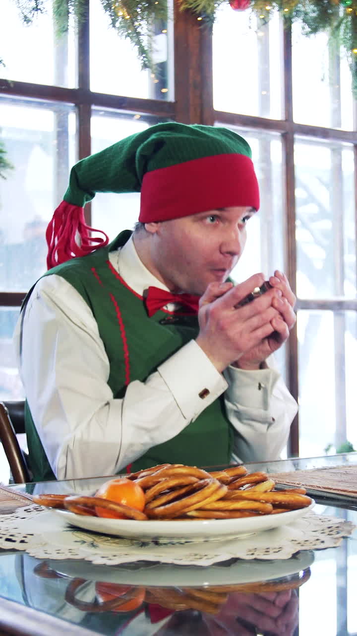Elf male with cup sitting at table on Christmas background. Funny elf in green costume gets up from a table. Vertical video