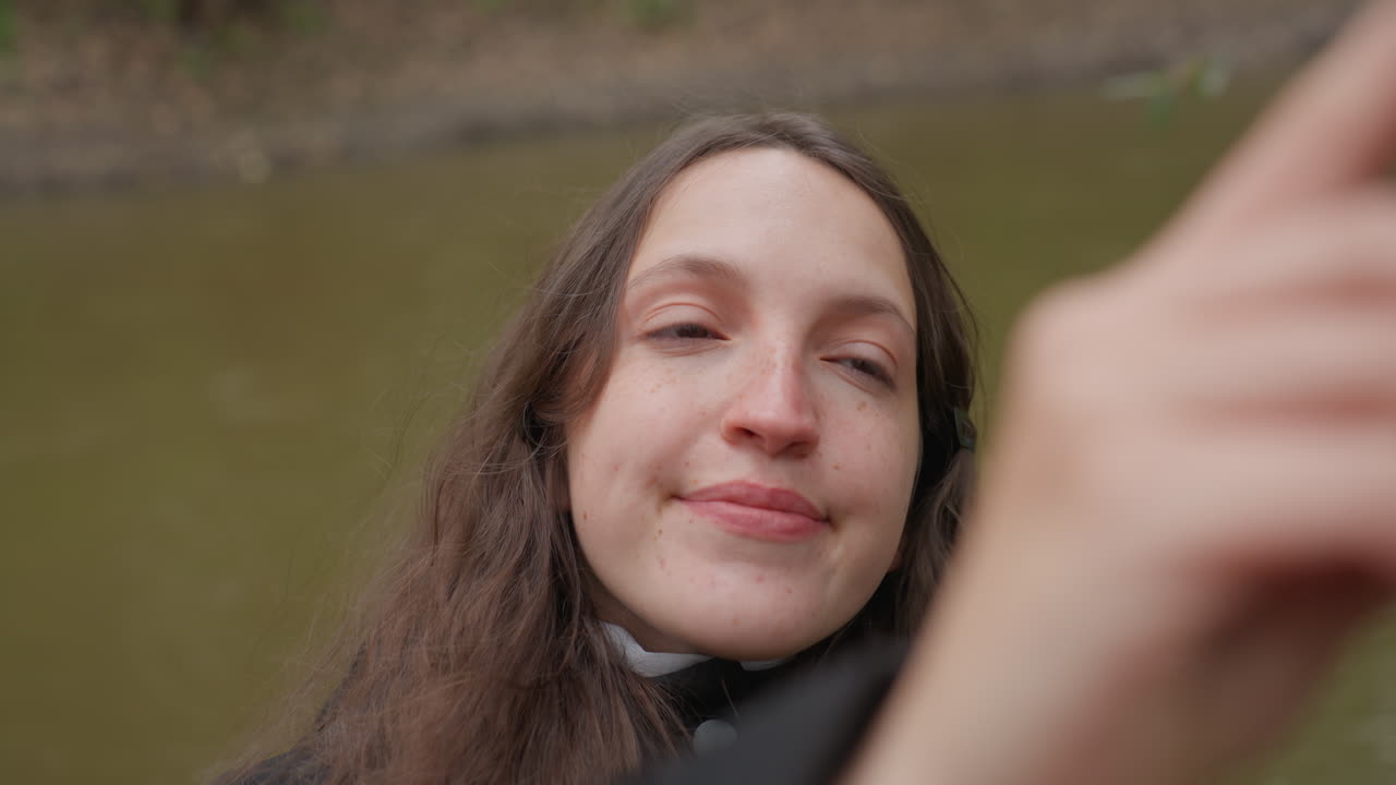Nainen joen rannalla ottaa selfietä, pysähtyy rauhallisella kävelyllä puun ja mutaisen rantatörmän vieressä. Pitkät, lainehtivat hiukset tungettu hupullisen takin sisään, kädessä pidettävä puhelin luo avoimia muotokuvia, pehmeä hymy ja lämmin.