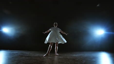 zoom camera moves around the stage with software and smoke. girl ballerina dancing in a white dress spinning plastic while performing pirouettes and rotations experiencing emotions and dramatic atmosphere.